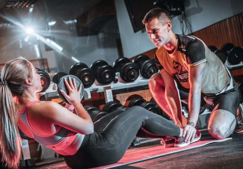 A personal trainer assists a woman doing sit-ups with a medicine ball in a gym setting.